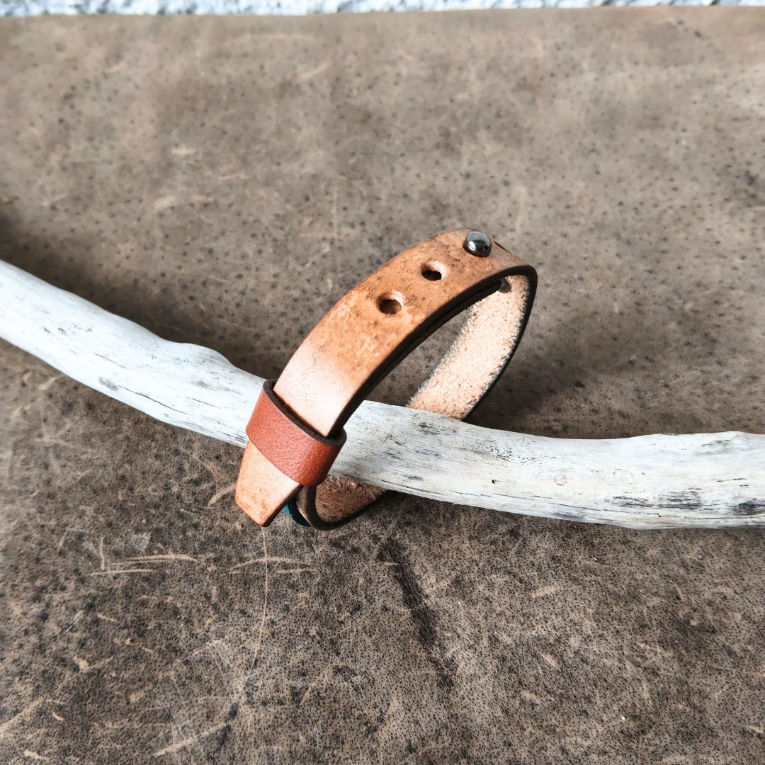 A handmade leather cuff bracelet in a warm tan-brown color, resting on a white driftwood branch over a textured brown surface.