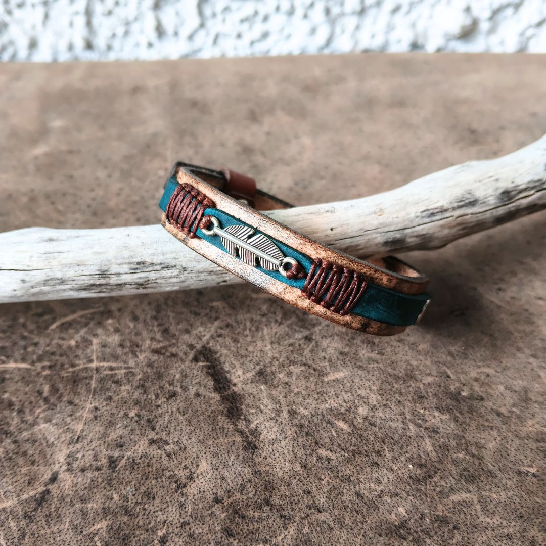 A handmade leather cuff bracelet with a turquoise blue inlay, brown braided cord accents, and a silver feather charm, resting on a piece of driftwood over a textured brown surface.