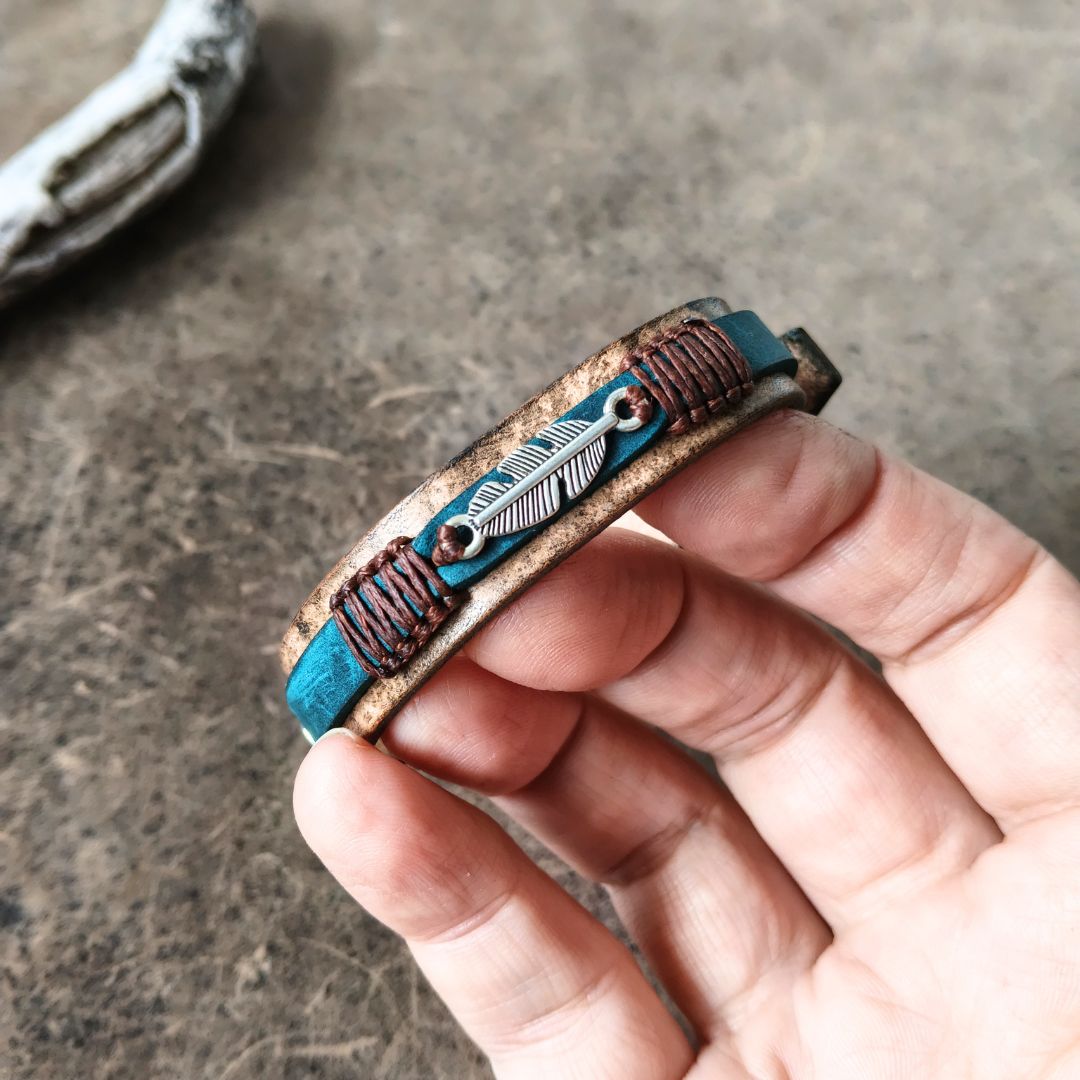 A hand holding a handmade leather cuff bracelet with turquoise blue inlay, copper wire wrapping, and a silver feather charm, set against a textured gray background.