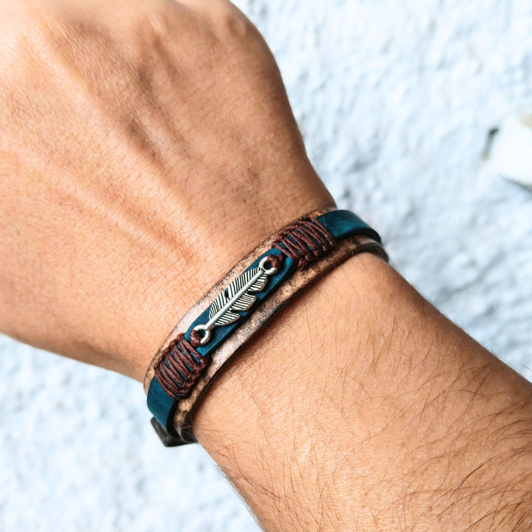 A close-up of a person's wrist wearing a multi-layered leather bracelet. The bracelet features a dark teal blue leather band, a brown leather band with dark brown stitching, and a central silver-toned feather charm .