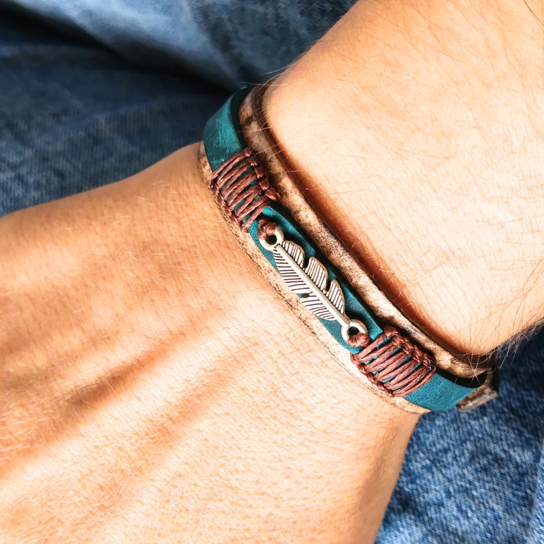 A close-up of a person's wrist wearing a tan leather bracelet. The bracelet features a teal leather band, a brown braided cord, and a central silver-toned feather.