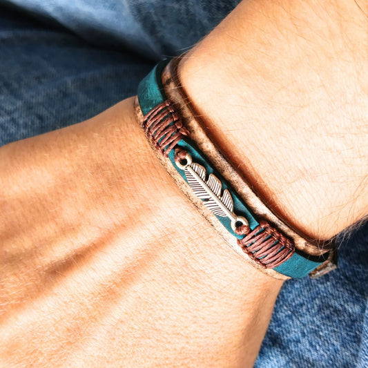 A close-up of a person's wrist wearing a tan leather bracelet. The bracelet features a teal leather band, a brown braided cord, and a central silver-toned feather.
