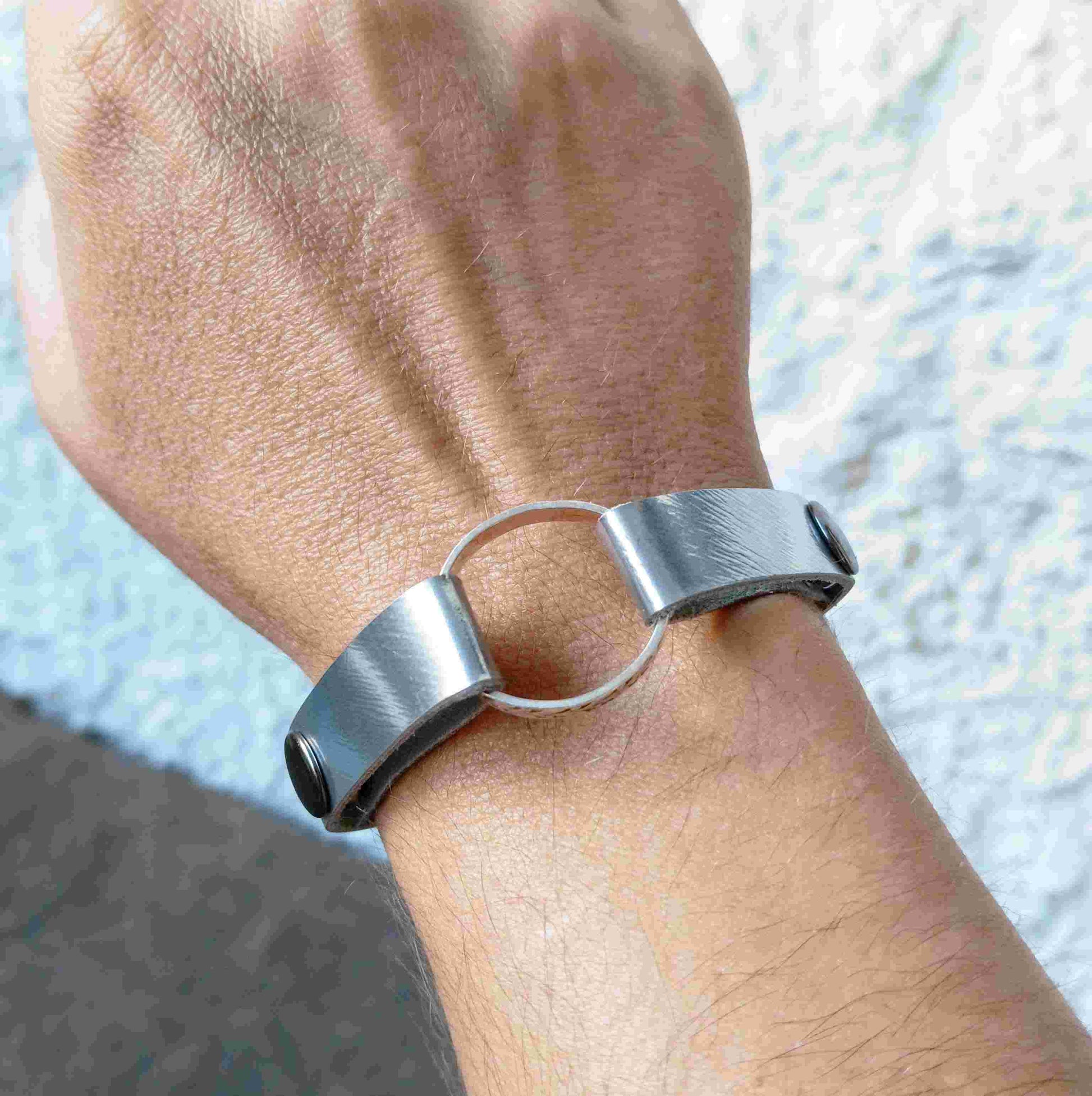 A close-up of a person's wrist wearing a minimalist muted silver leather cuff bracelet with a central circular ring.