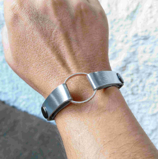 A close-up of a person's wrist wearing a minimalist muted silver leather cuff bracelet with a central circular ring.
