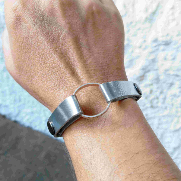 A close-up of a person's wrist wearing a minimalist muted silver leather cuff bracelet with a central circular ring.