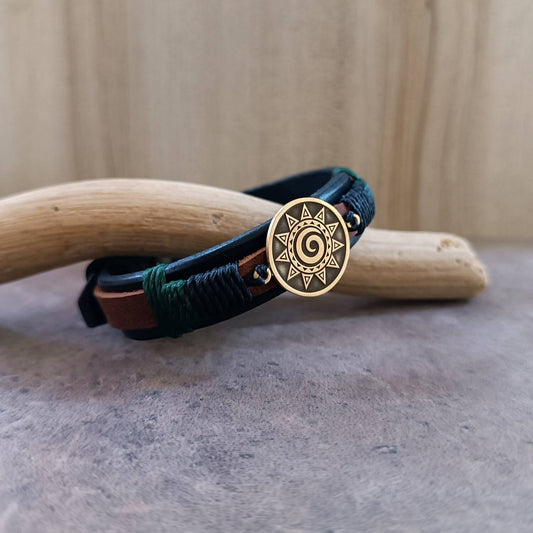 Unisex black leather watch-band style bracelet with thin brown leather strip and brass Polynesian Koru spiral sun pendant.
