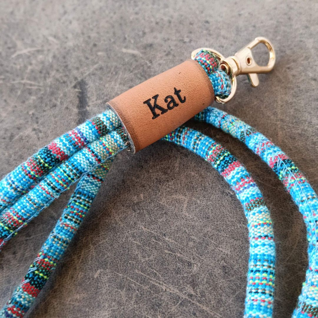 A close-up of a personalized woven rope keychain or bag strap with a tan leather cylinder engraved with the name 'Kat' and a gold-tone metal clasp, set against a textured gray background.