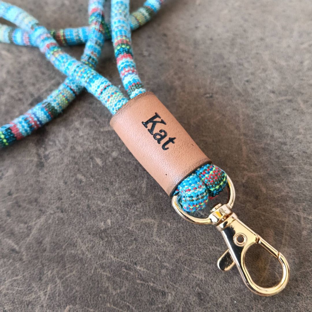 A light blue and white braided rope keychain with a brown leather tag engraved with the name 'Kat' and silver metal key rings and clasp, displayed on a dark textured surface.