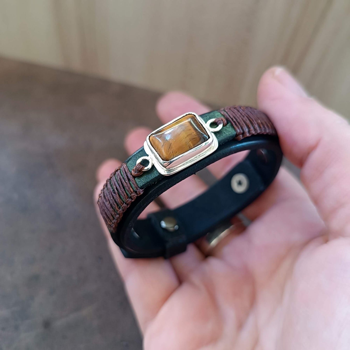 Hand holding a black leather bracelet with a tiger eye stone against a neutral background