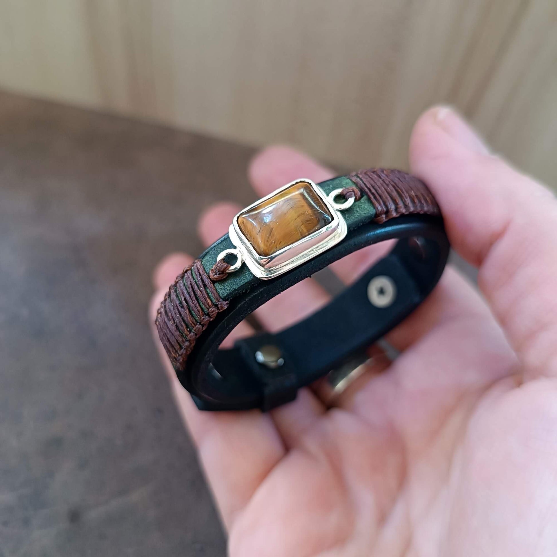 Hand holding a black leather bracelet with a tiger eye stone against a neutral background