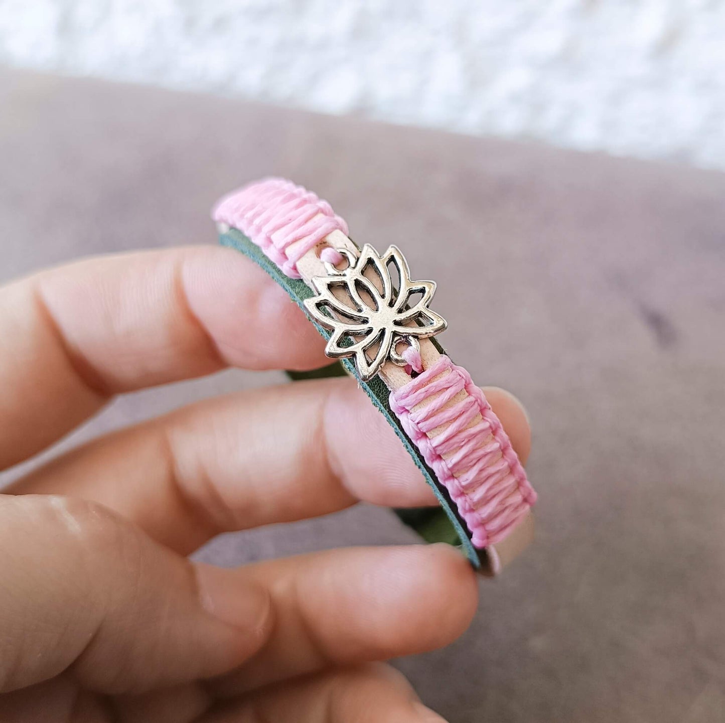 A handmade pink braided leather bracelet with a silver lotus flower charm, held in a person's hand against a neutral background.