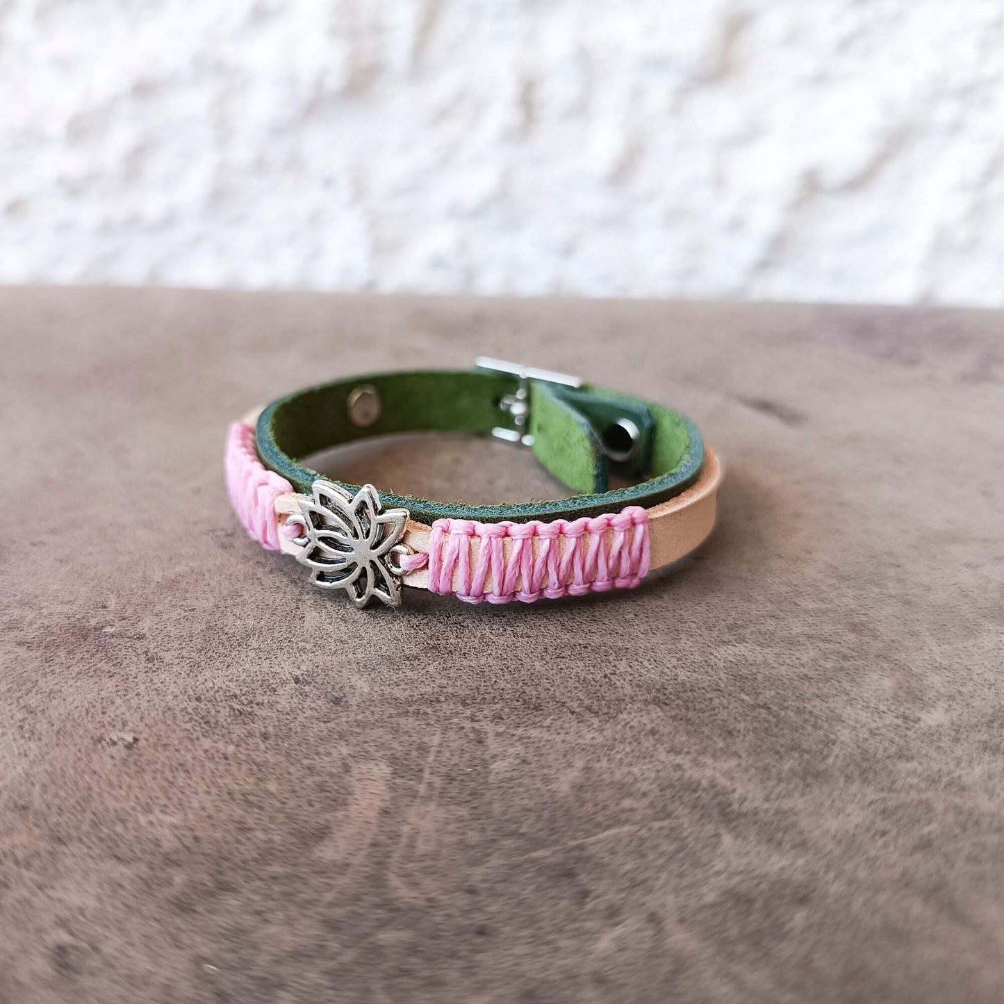 A handmade leather bracelet with a green inner lining, pink braided cord wrapping, a silver-toned metal lotus flower charm, and a tan leather accent band, resting on a textured brown surface.