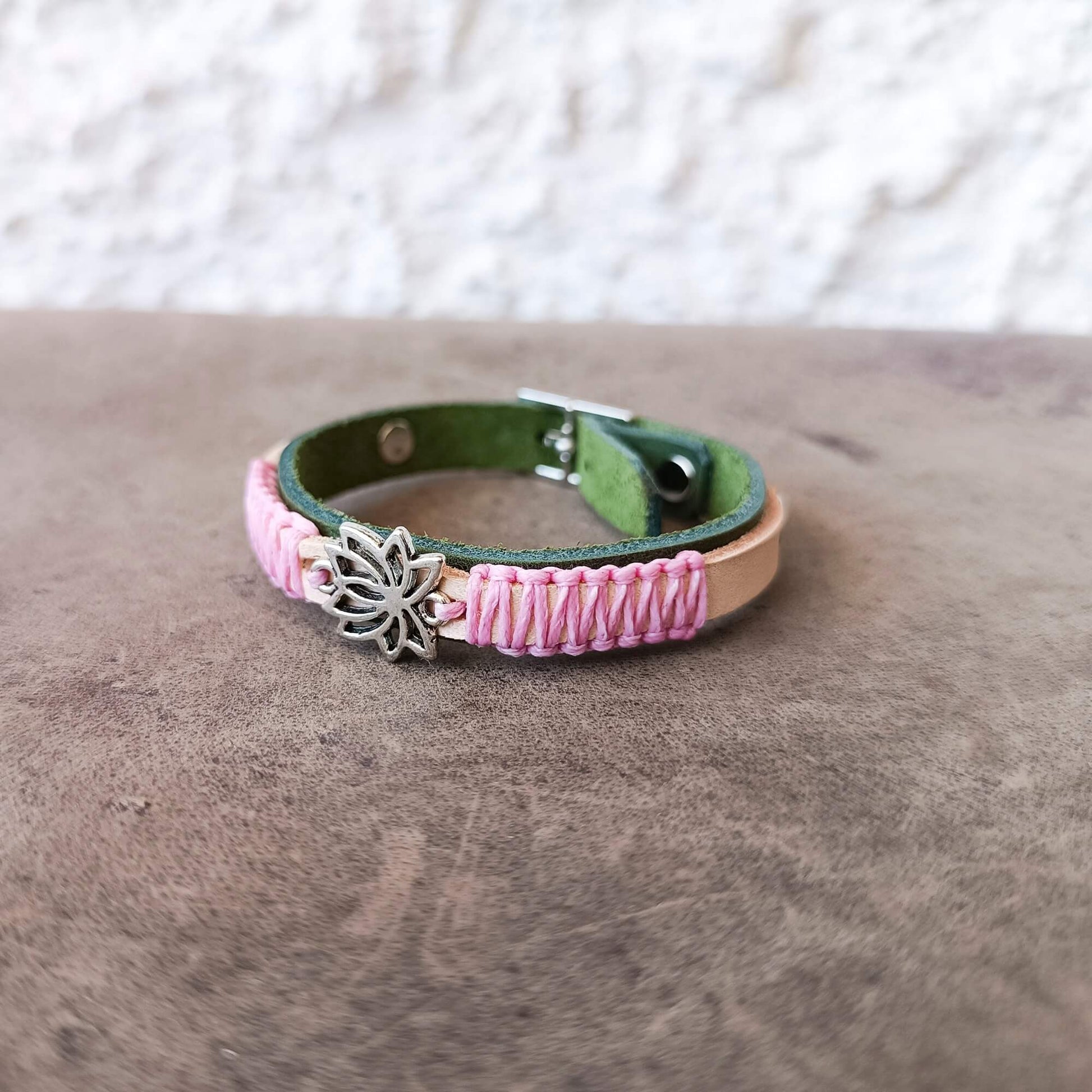 A handmade leather bracelet with a green inner lining, pink braided cord wrapping, a silver-toned metal lotus flower charm, and a tan leather accent band, resting on a textured brown surface.