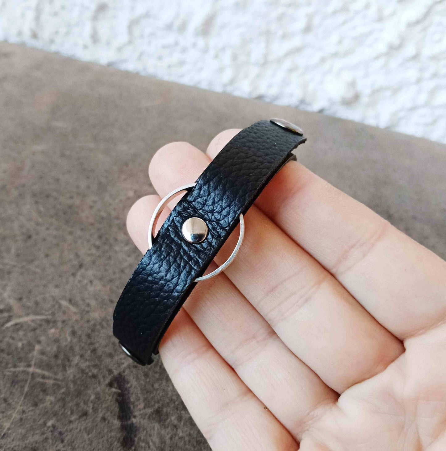 A close-up of a hand holding a black textured leather ring holder bracelet with a silver metal ring and stud closure, set against a neutral background.