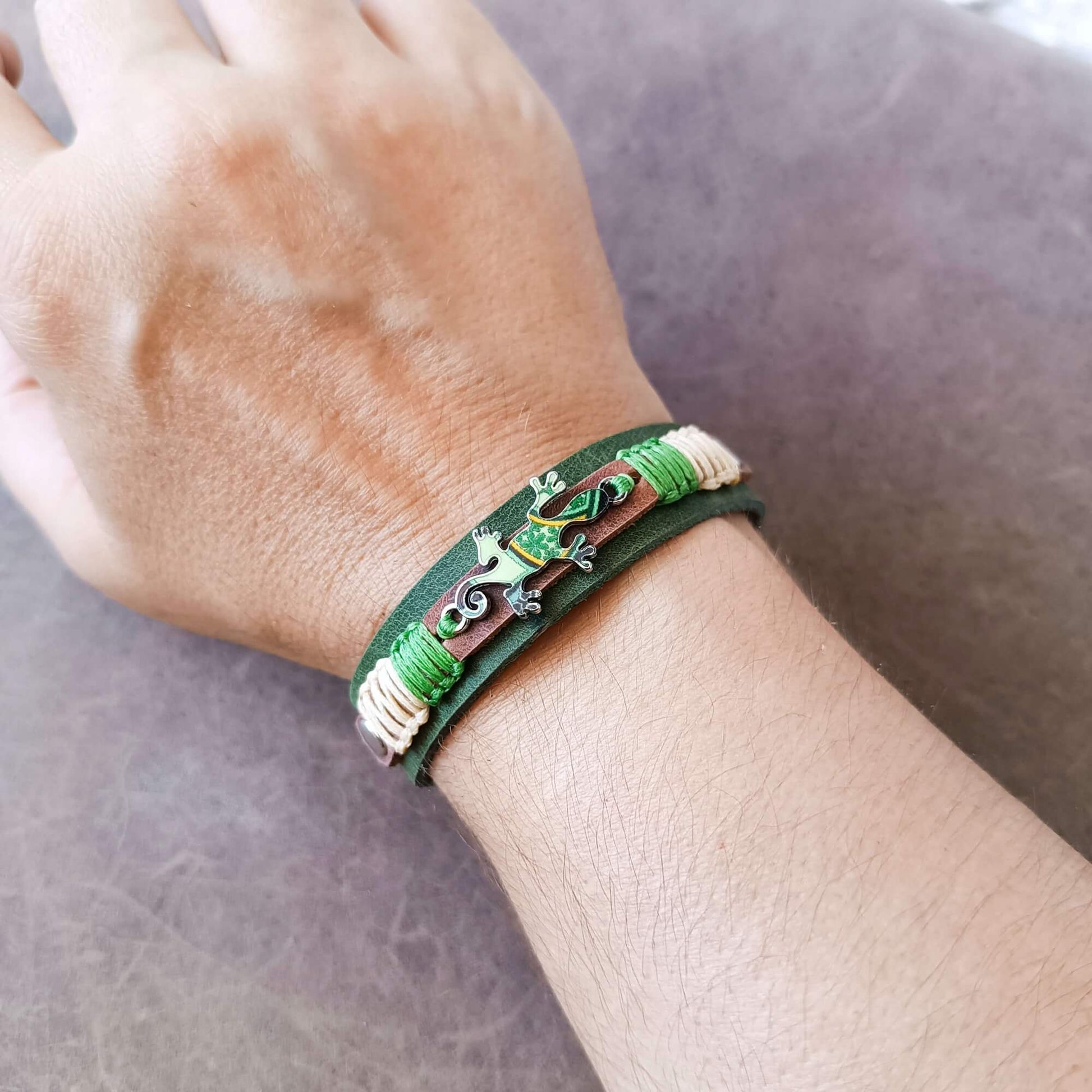A close-up of a person's wrist wearing a handmade leather bracelet. The bracelet features a dark green leather band accented with green and cream braided cord, a small silver-toned Gecko charm, and a decorative brown and green scroll element.